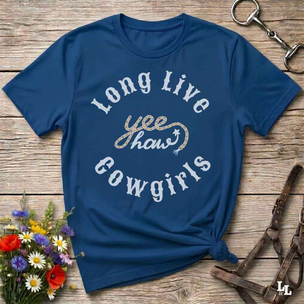 A navy blue graphic t-shirt with 'Long Live yee haw cowgirls' printed on the front, styled in a flat-lay on a rustic wooden background with flowers and horse tack.