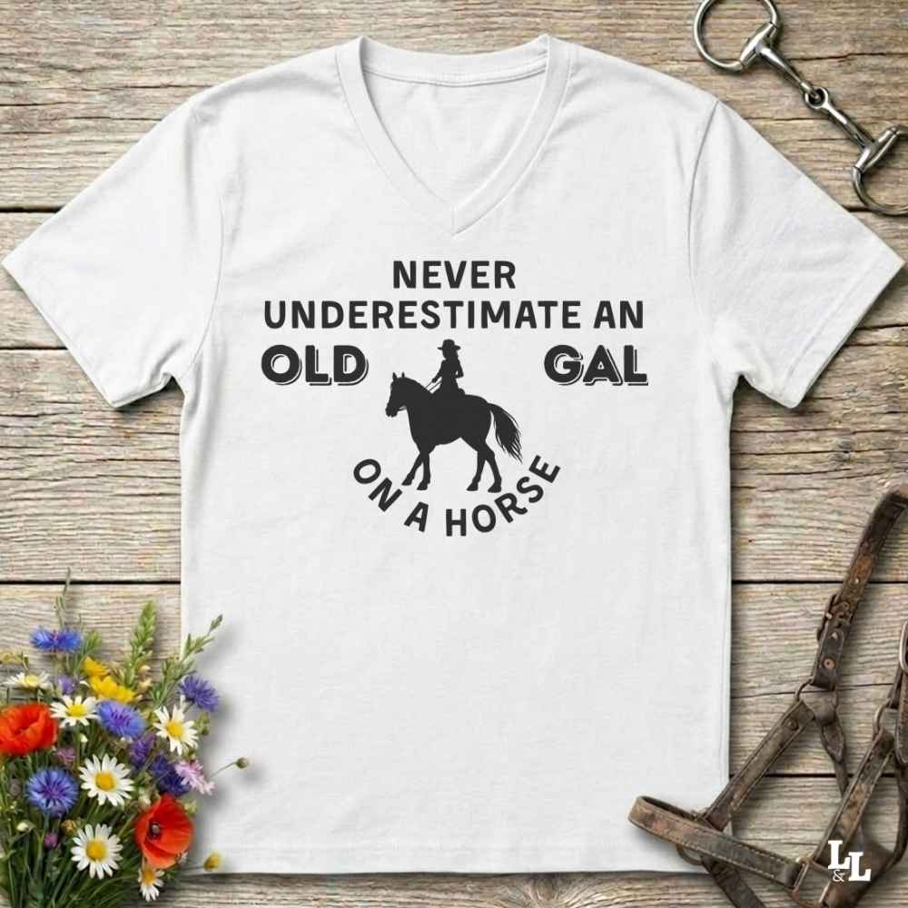 A white V-neck t-shirt with a black graphic print reading 'NEVER UNDERESTIMATE AN OLD GAL ON A HORSE' with a silhouette of a rider on a horse, displayed flat on a rustic wooden background with equestrian accessories and flowers.