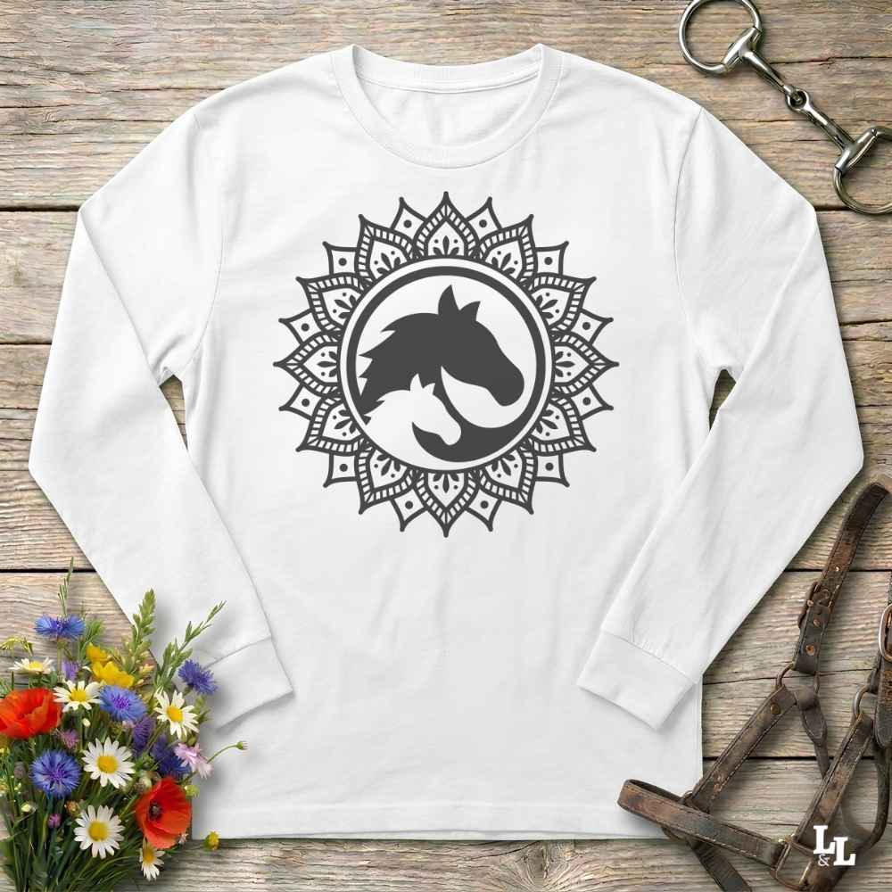 A long-sleeve crewneck shirt in white red, featuring a black graphic of a horse and cat silhouette inside a circular mandala design. The shirt is displayed flat on a rustic wooden background with equestrian tack and a small bouquet of flowers.