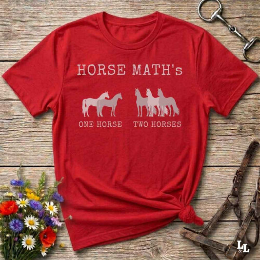 A red short-sleeve graphic T-shirt laid flat on a rustic wooden surface, with white text reading 'HORSE MATH'S' and horse silhouettes above the phrases 'ONE HORSE' and 'TWO HORSES.' Decorative flowers and horse tack are placed around the shirt as props.
