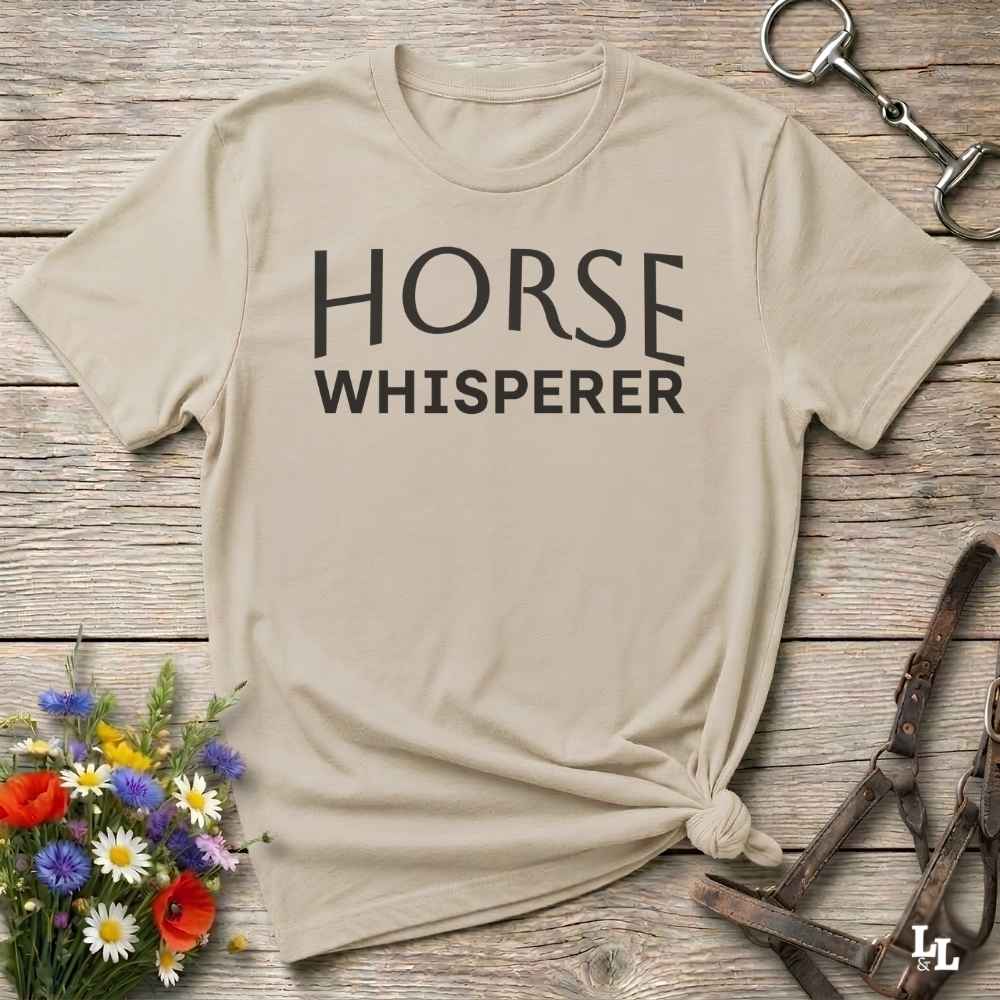 A beige graphic t-shirt with the words 'HORSE WHISPERER' printed in black across the chest, styled in a flat-lay on a rustic wooden background with a horse bit, leather bridle, and a small bouquet of wildflowers.