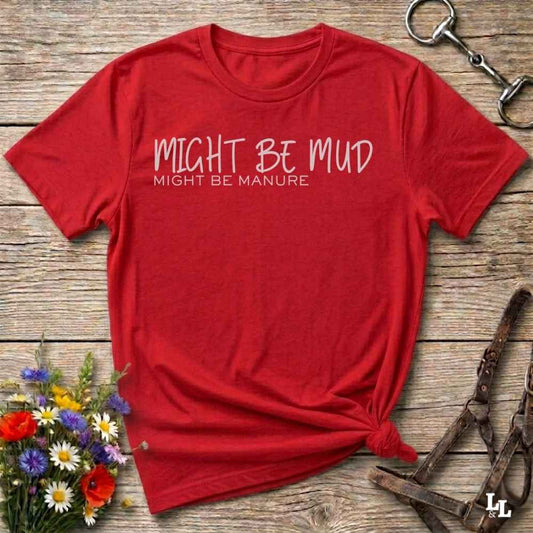 A red short-sleeve t-shirt with the text 'MIGHT BE MUD' and 'MIGHT BE MANURE' printed in white on the chest, styled flat on a rustic wooden background with equestrian accessories and flowers.