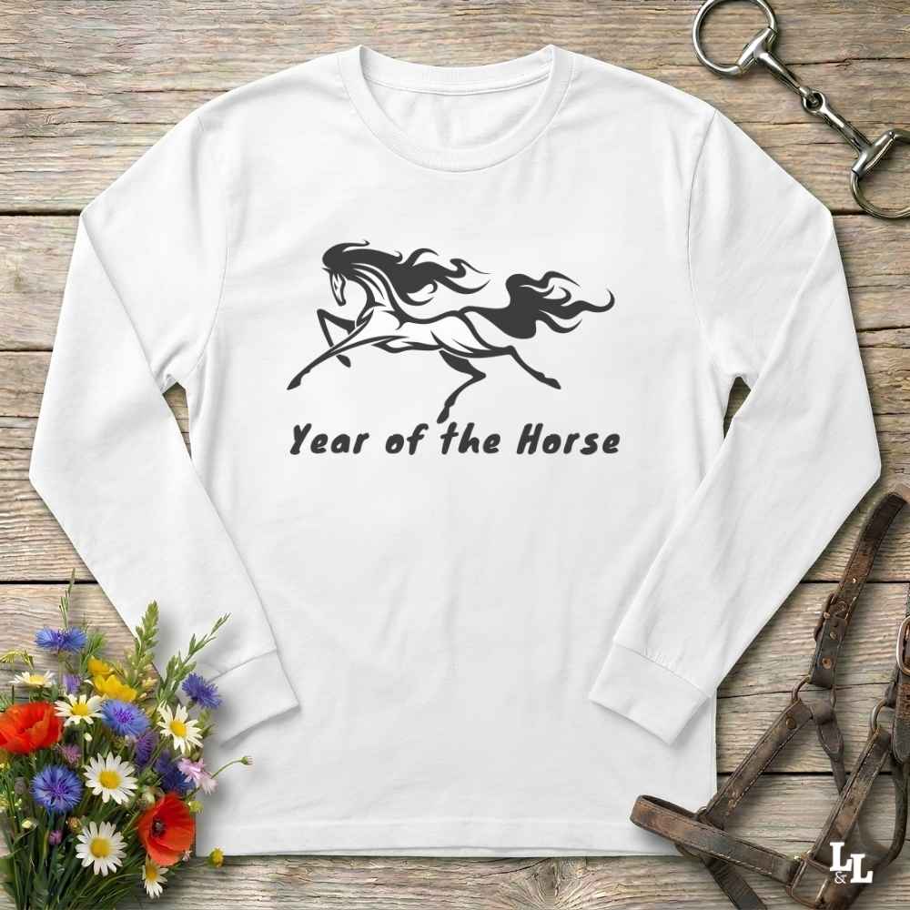 A white long-sleeve t-shirt with a black graphic of a rearing horse and the text 'Year of the Horse' printed on the chest, displayed flat on a rustic wooden background with equestrian accessories and flowers.