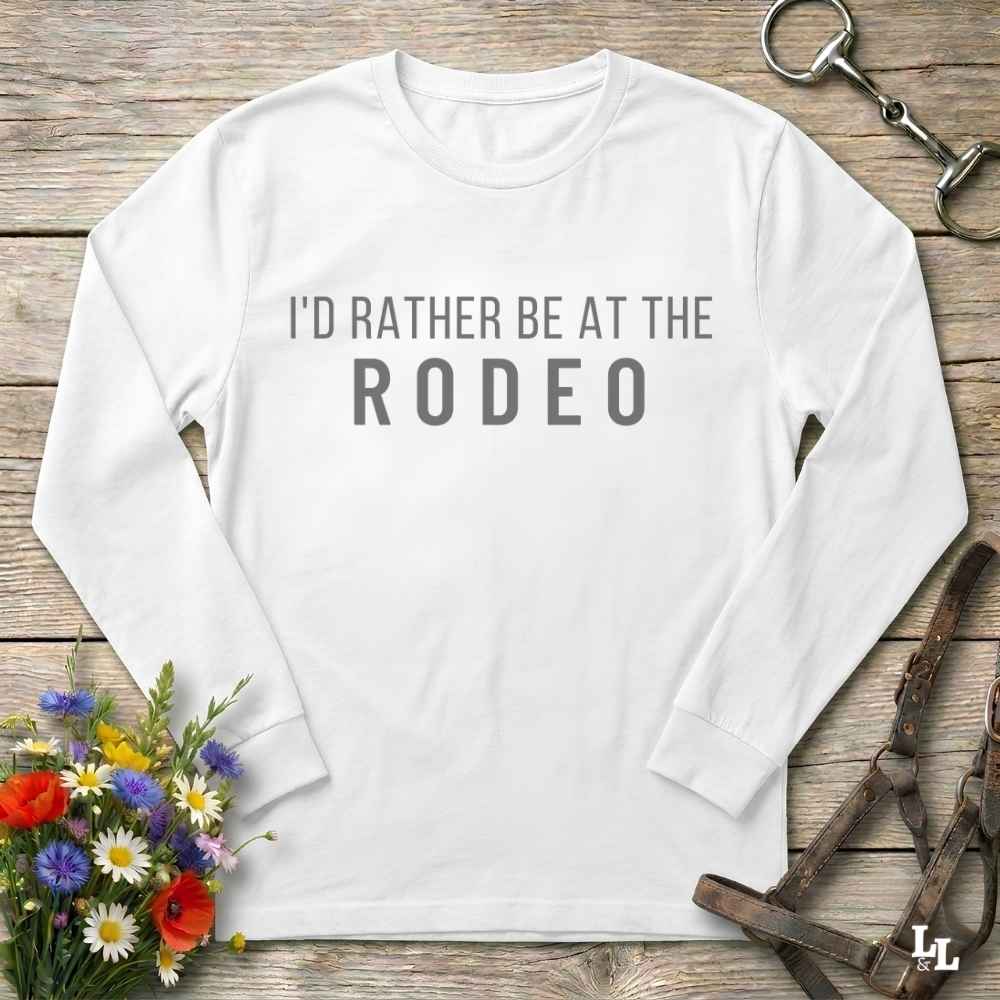 A white long-sleeve t-shirt with the text 'I'D RATHER BE AT THE RODEO' printed in gray across the chest, styled in a flat-lay on a rustic wooden background with equestrian tack and flowers.
