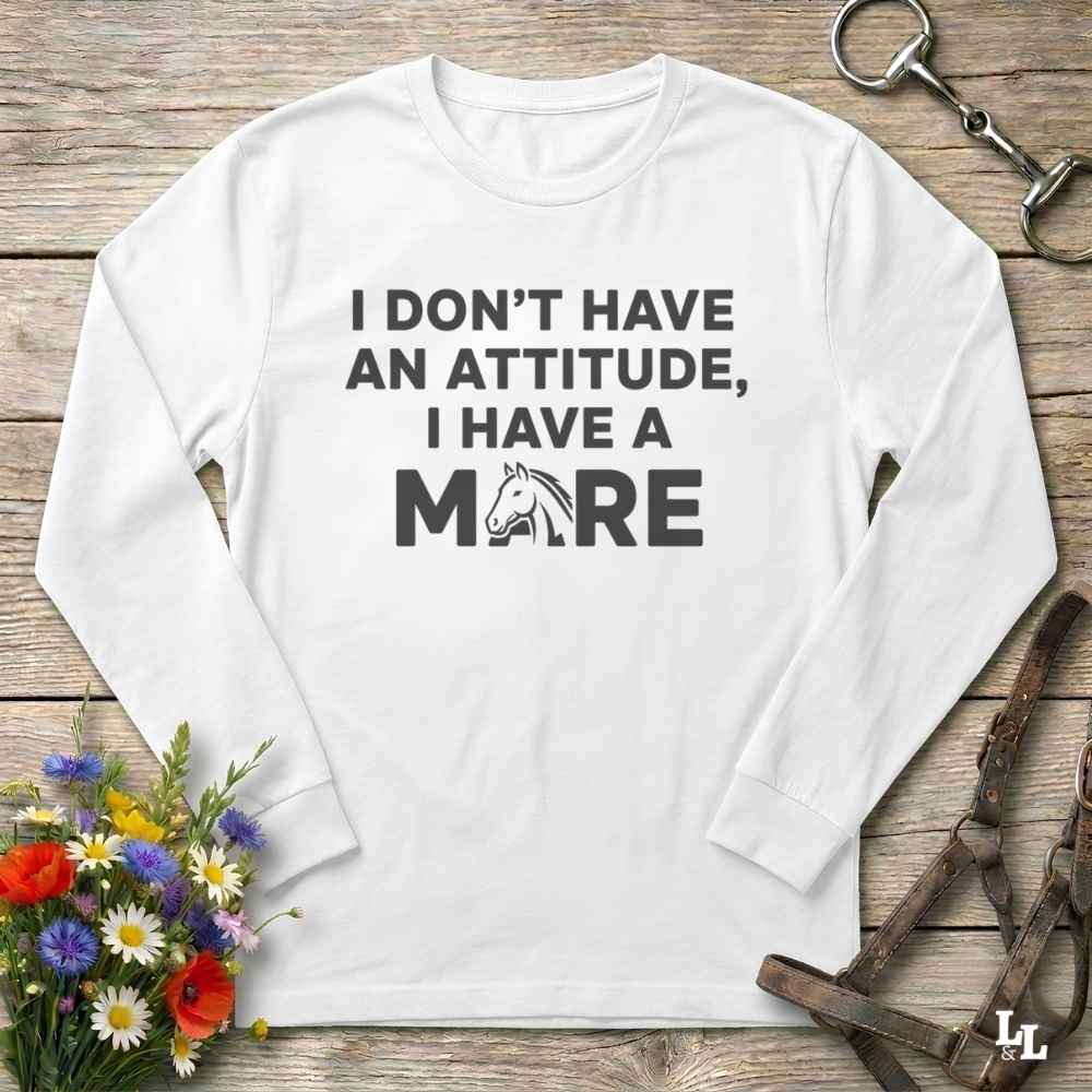 A white long-sleeve t-shirt with a humorous equestrian graphic reading 'I DON'T HAVE AN ATTITUDE, I HAVE A MORE' with a horse head silhouette, displayed on a rustic wooden background with equestrian accessories and flowers.