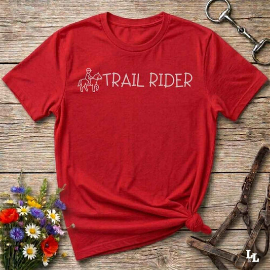 A red short-sleeve crewneck t-shirt with a white graphic reading 'TRAIL RIDER' and a small rider-and-horse outline, styled flat on a rustic wood surface with flowers, a halter, and a horse bit nearby.
