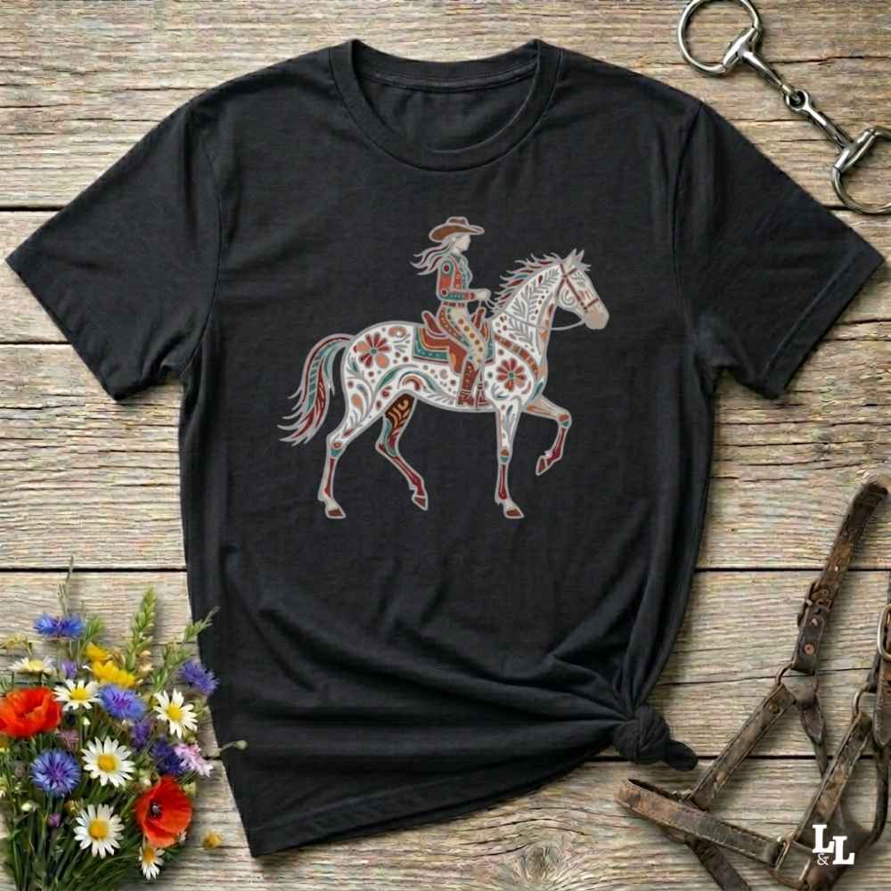 Black t-shirt with a colorful horse and rider graphic on a wooden surface with floral and equestrian-themed accessories.