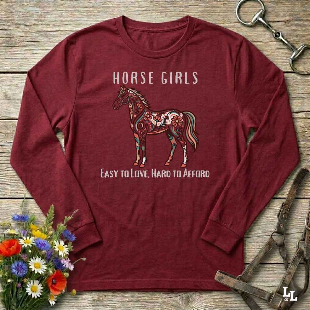 Maroon long-sleeve shirt with horse design and text on a wooden surface with flowers and equestrian equipment.