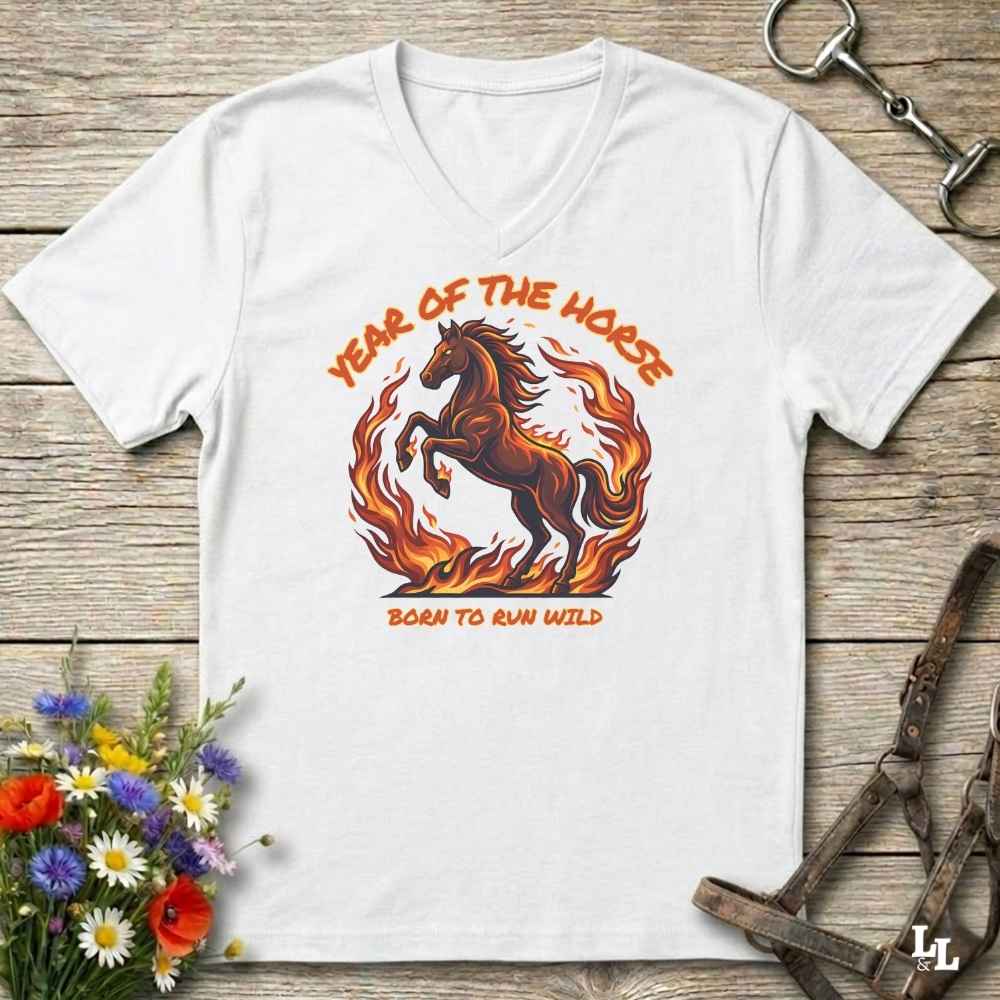 A white V-neck t-shirt laid flat on a rustic wooden surface, featuring a graphic of a rearing horse surrounded by flames with the text 'YEAR OF THE HORSE' and 'BORN TO RUN WILD'. The shirt is styled with equestrian props including a horse bit, leather tack, and a small bouquet of wildflowers.