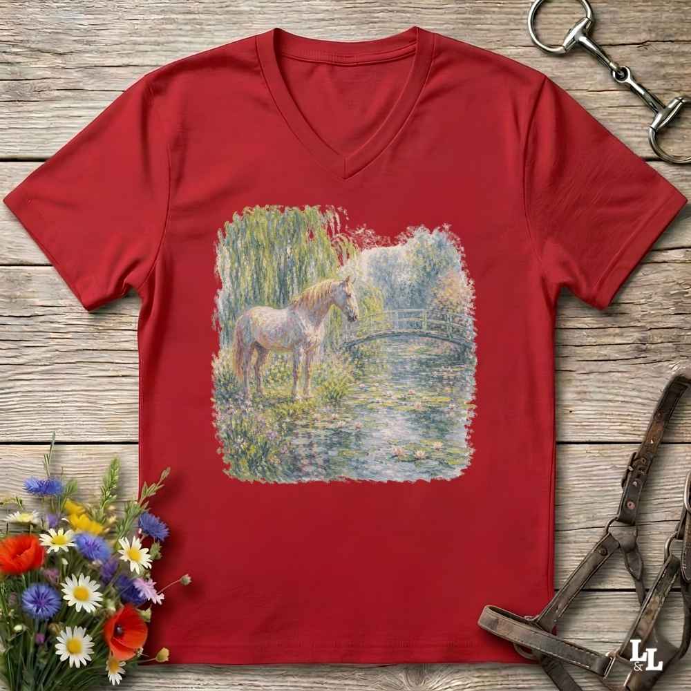 A red V-neck t-shirt laid flat on a rustic wooden surface, featuring a colorful watercolor-style print of a horse standing by a pond. The shirt is styled with equestrian accessories (reins and bit) and a small bouquet of wildflowers.