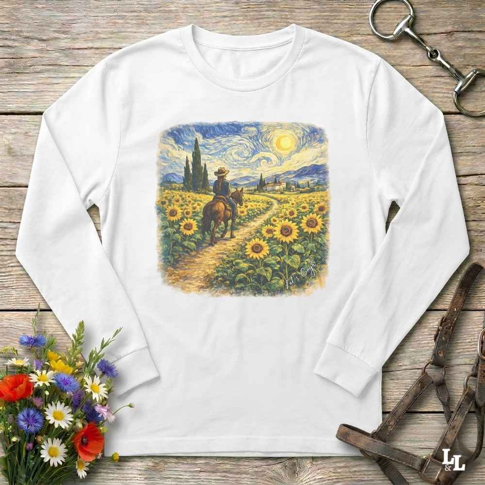 A white long-sleeve t-shirt with a colorful graphic print of a cowboy riding through a sunflower field in a Van Gogh-inspired style. The shirt is displayed flat on a rustic wooden background with decorative floral and equestrian props.