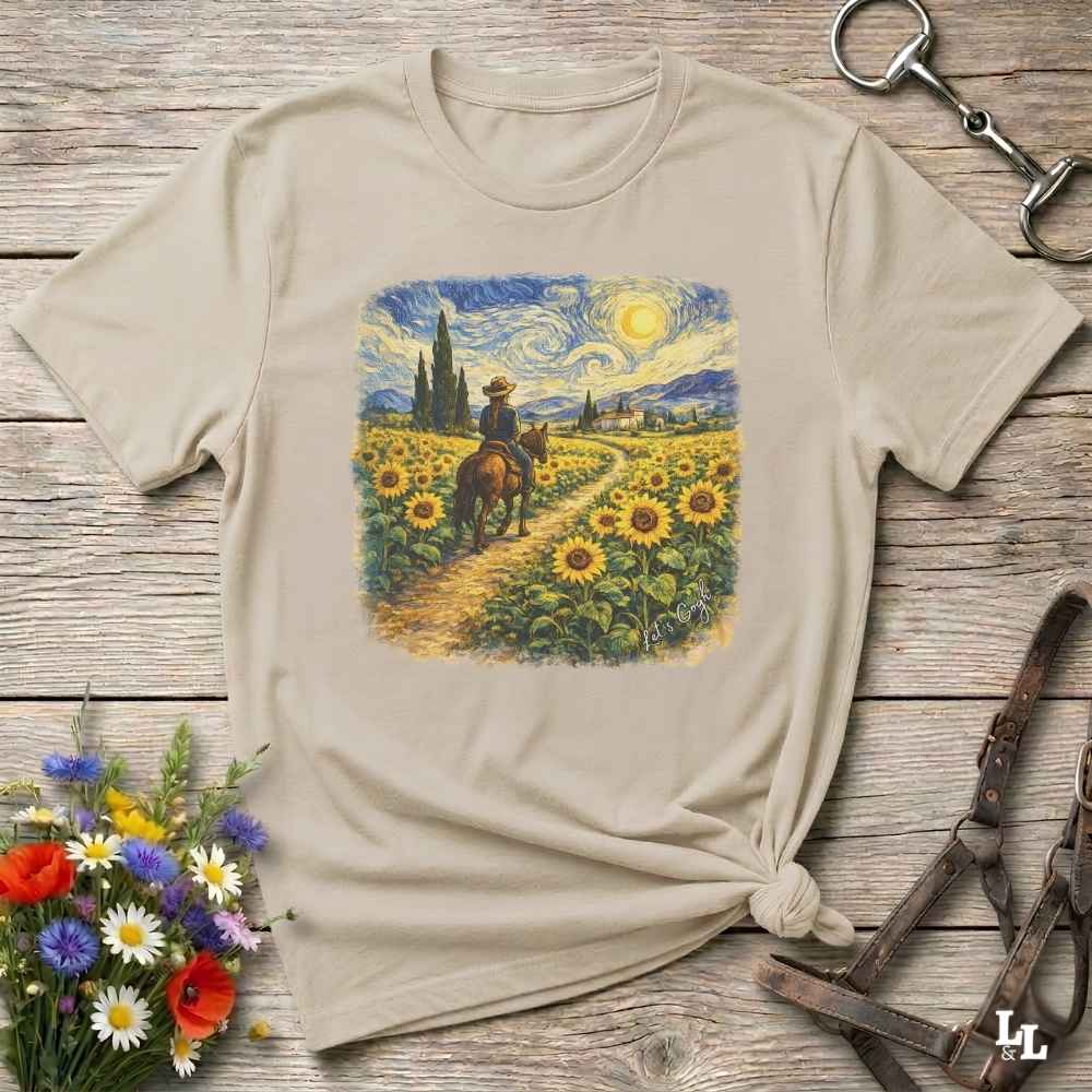 A beige short-sleeve t-shirt with a colorful graphic print of a cowboy riding through a sunflower field, styled on a rustic wooden background with a horse bit and a small bouquet of wildflowers.