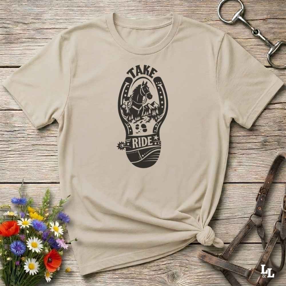 A beige short-sleeve t-shirt with a black graphic print of a horse and the words 'TAKE RIDE' inside a horseshoe design, styled on a rustic wooden background with equestrian accessories and flowers.