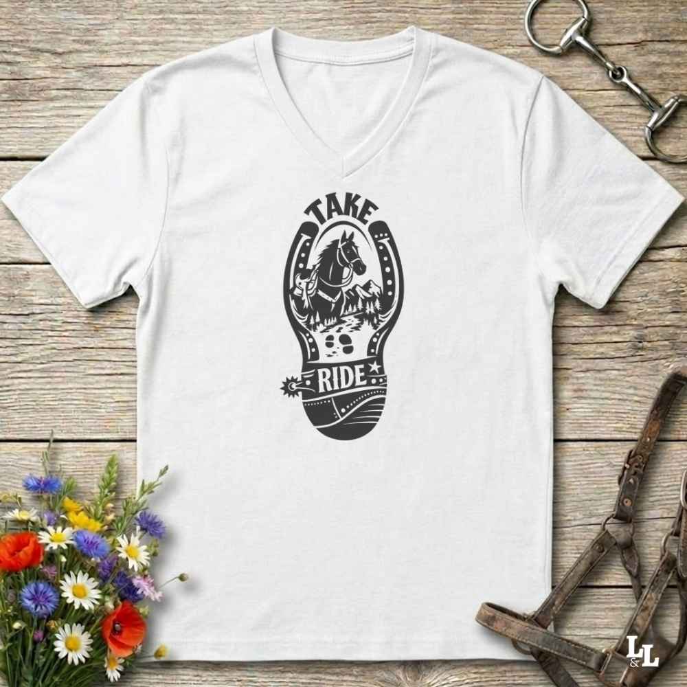 A white V-neck t-shirt with a light gray graphic of a horse inside a horseshoe and the words 'TAKE RIDE', displayed on a rustic wooden background with equestrian accessories and flowers.