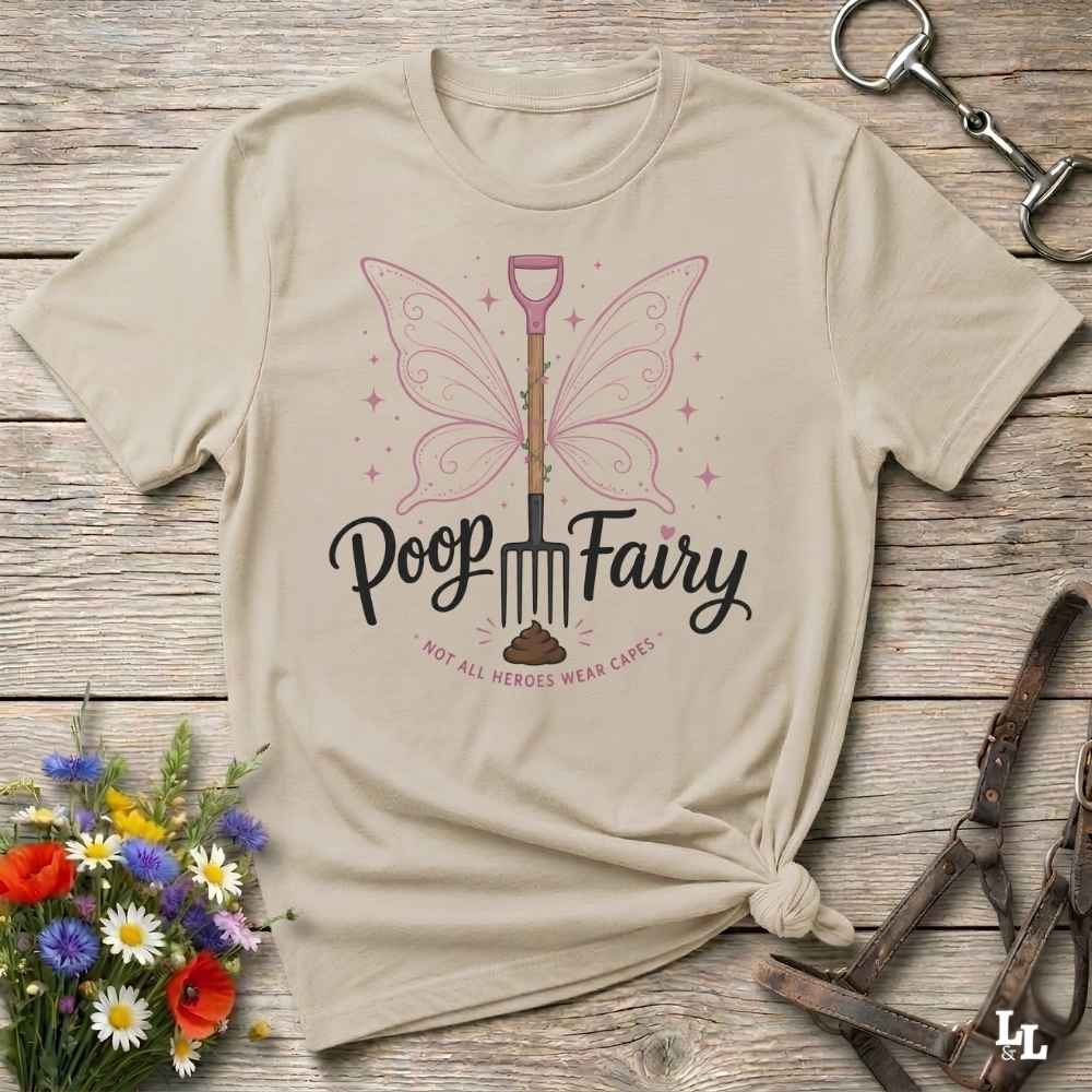 A beige graphic t-shirt laid flat on a rustic wooden surface, styled with equestrian accessories and a small bouquet of flowers. The shirt features a graphic of a fairy-winged pitchfork with the text 'Poop Fairy' and the phrase 'NOT ALL HEROES WEAR CAPES'.