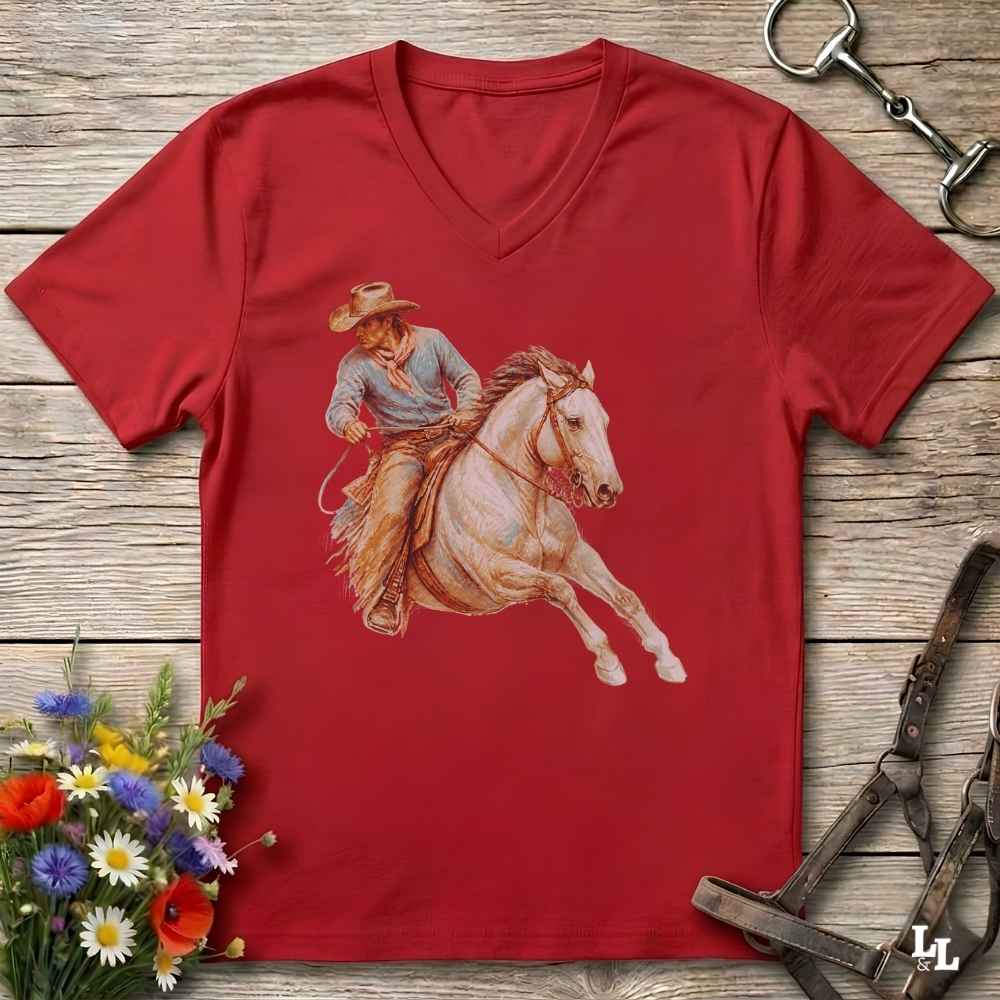 A red V-neck t-shirt featuring a western cowboy riding a white horse graphic, displayed flat on a rustic wooden background with equestrian and floral props.