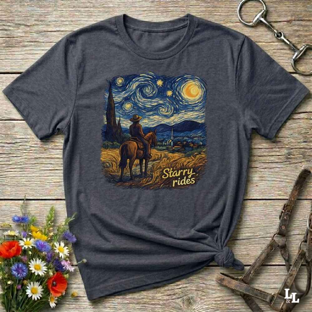 A dark heather gray graphic t-shirt laid flat on a rustic wooden surface, styled with a bouquet of flowers, a horse bit, and a leather halter. The t-shirt features a central graphic print in a Van Gogh-inspired style showing a cowboy on a horse under a swirling starry night sky, with the text 'Starry rides'.