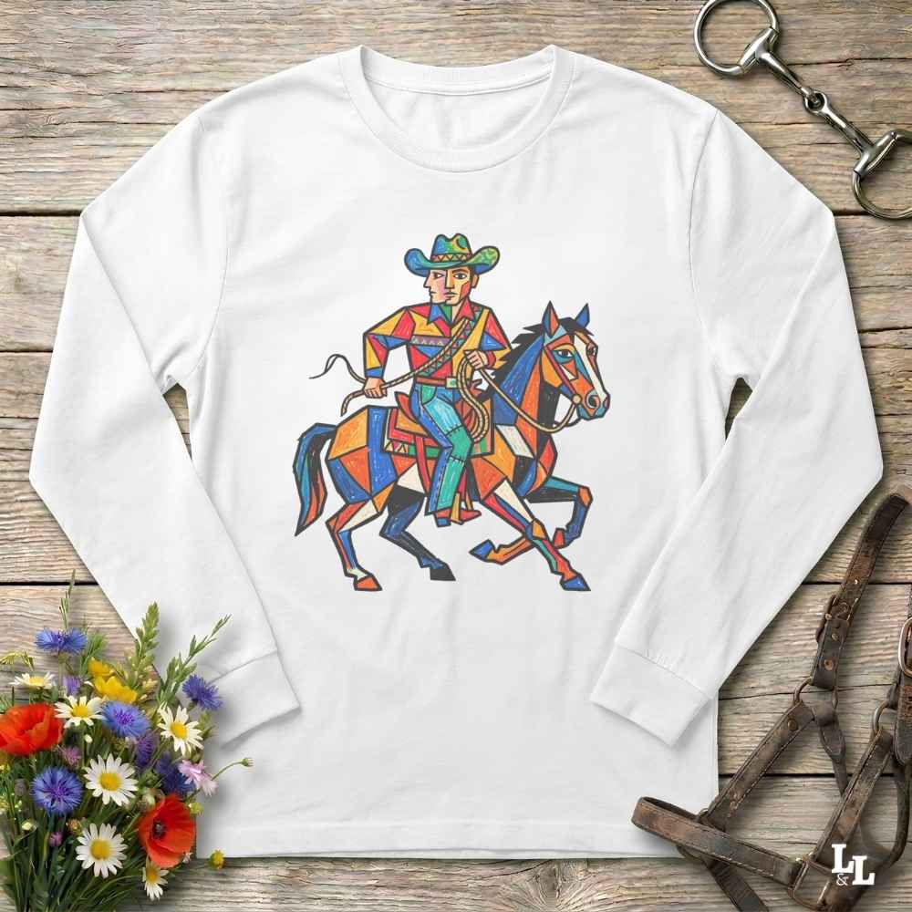 A white long-sleeve t-shirt with a colorful geometric graphic of a cowboy riding a horse, displayed on a rustic wooden background with decorative flowers and leather tack.