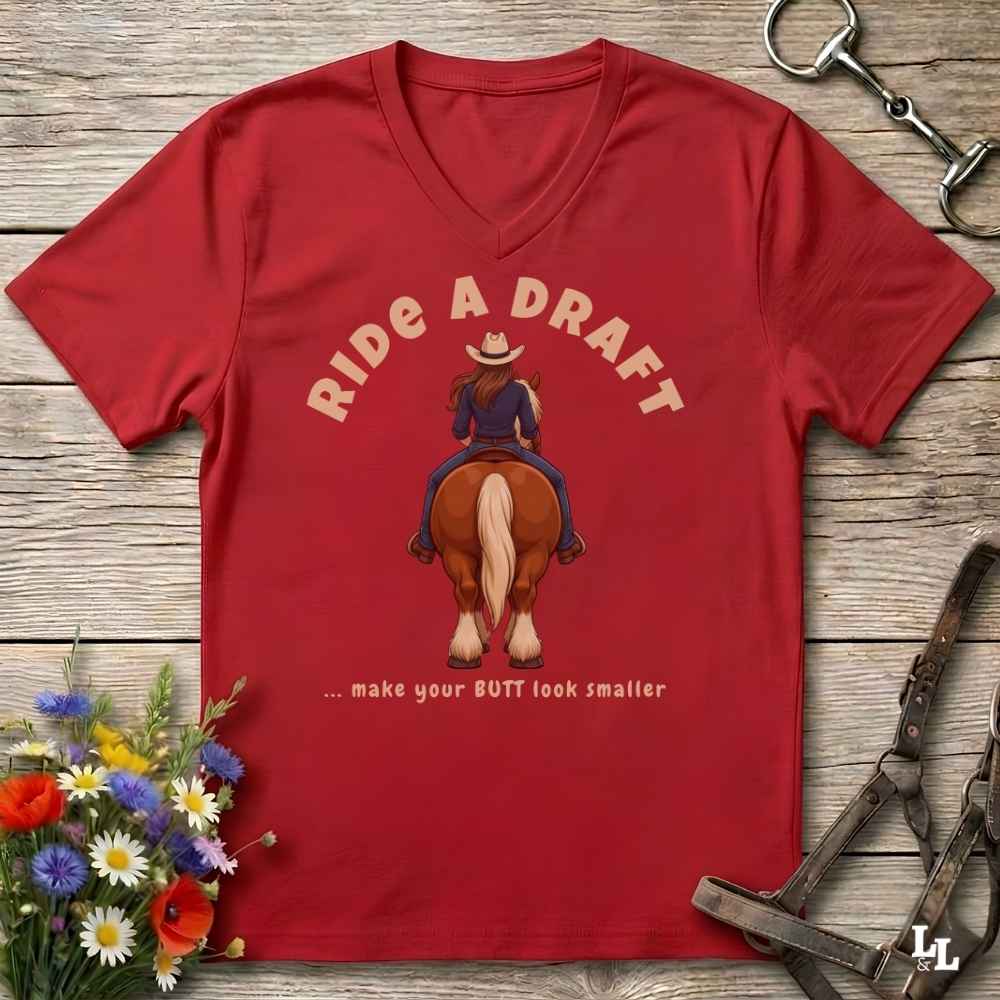 A red V-neck t-shirt laid flat on a rustic wooden surface, featuring a graphic of a woman riding a brown horse from behind with the text 'RIDE A DRAFT .
