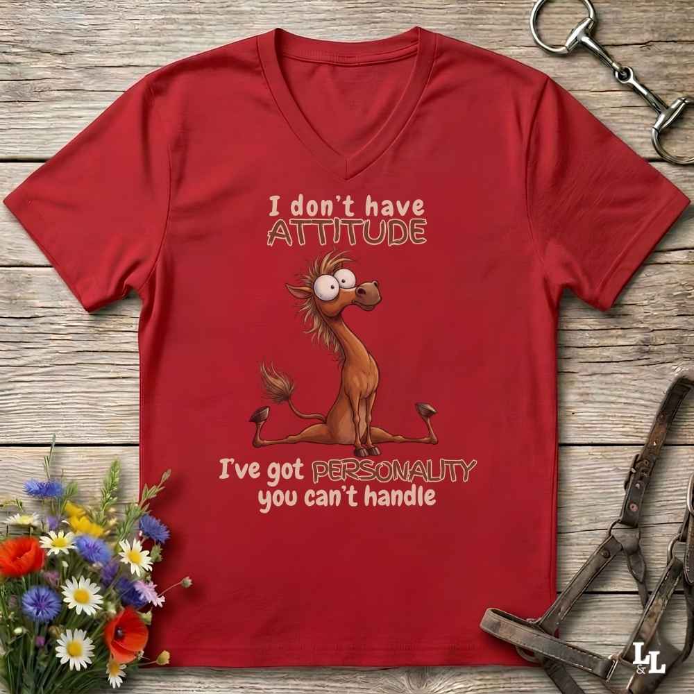 A red V-neck t-shirt with a humorous cartoon horse graphic and the text 'I don't have ATTITUDE I've got PERSONALITY you can't handle', displayed on a rustic wooden background with horse tack and flowers.