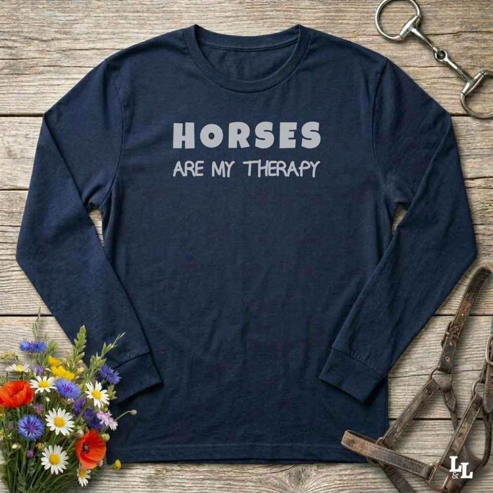 A navy blue long-sleeve t-shirt with the text 'HORSES ARE MY THERAPY' printed in white on the chest, styled in a flat-lay on a rustic wooden background with equestrian and floral props.