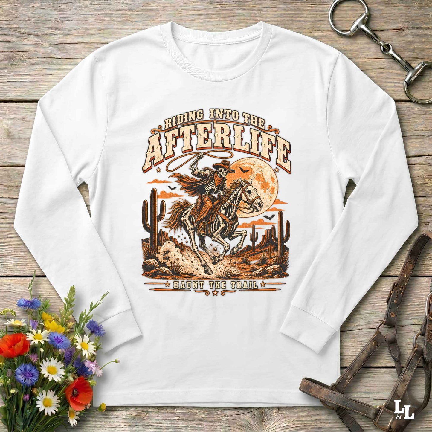 A white long-sleeve t-shirt with a western-themed graphic print, laid flat on a rustic wooden background with decorative props including flowers, a horse bit, and a leather bridle.