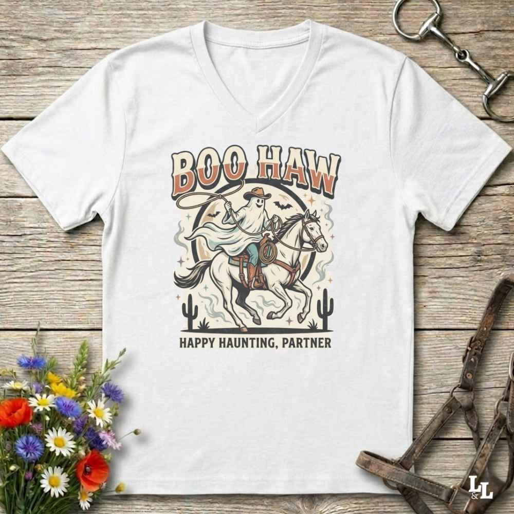 A white V-neck t-shirt with a western-themed 'Boo Haw Happy Haunting, Partner' graphic, displayed flat on a rustic wooden background with decorative horse tack and flowers.