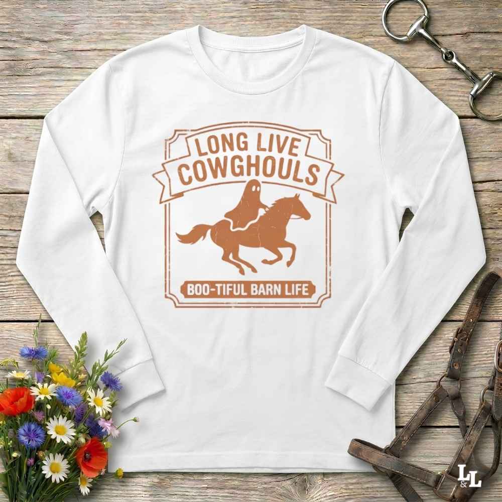 A white long-sleeve crewneck t-shirt with a brown graphic print of a ghost riding a horse and the text 'LONG LIVE COWGHOULS' and 'BOO-TIFUL BARN LIFE'. The shirt is displayed flat on a rustic wooden background with equestrian and floral props.