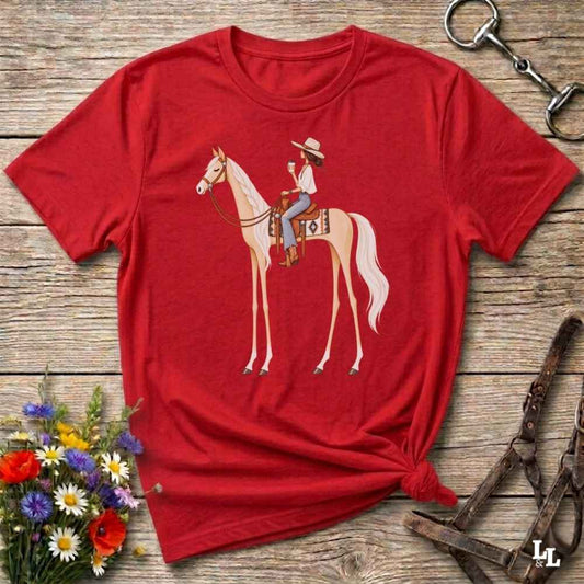 A red short-sleeve crewneck t-shirt with a graphic of a woman riding a light-colored horse, styled in a flat-lay on a rustic wooden background with equestrian accessories and flowers.