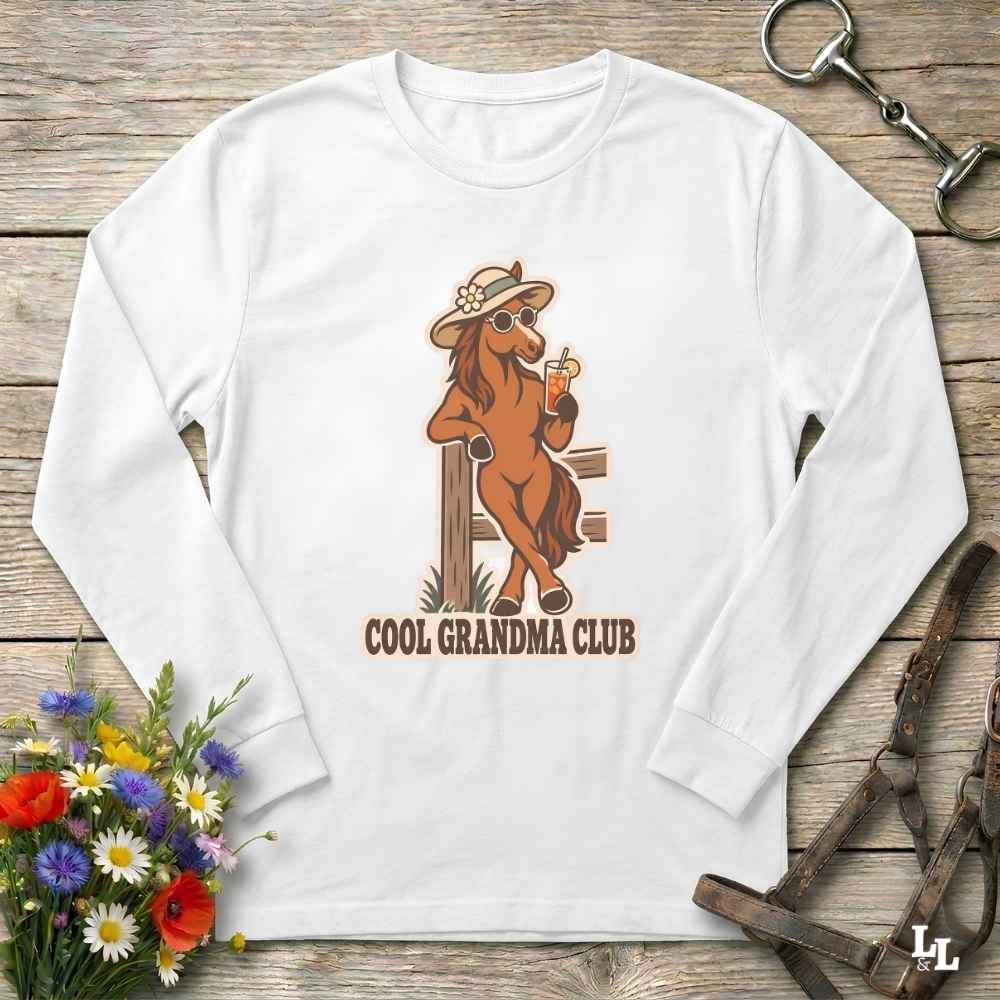 A white long-sleeve crewneck t-shirt with a graphic of a cool brown horse wearing a hat and sunglasses, holding a drink, with the text 'COOL GRANDMA CLUB' underneath. The shirt is displayed flat on a rustic wooden background with equestrian accessories and flowers.