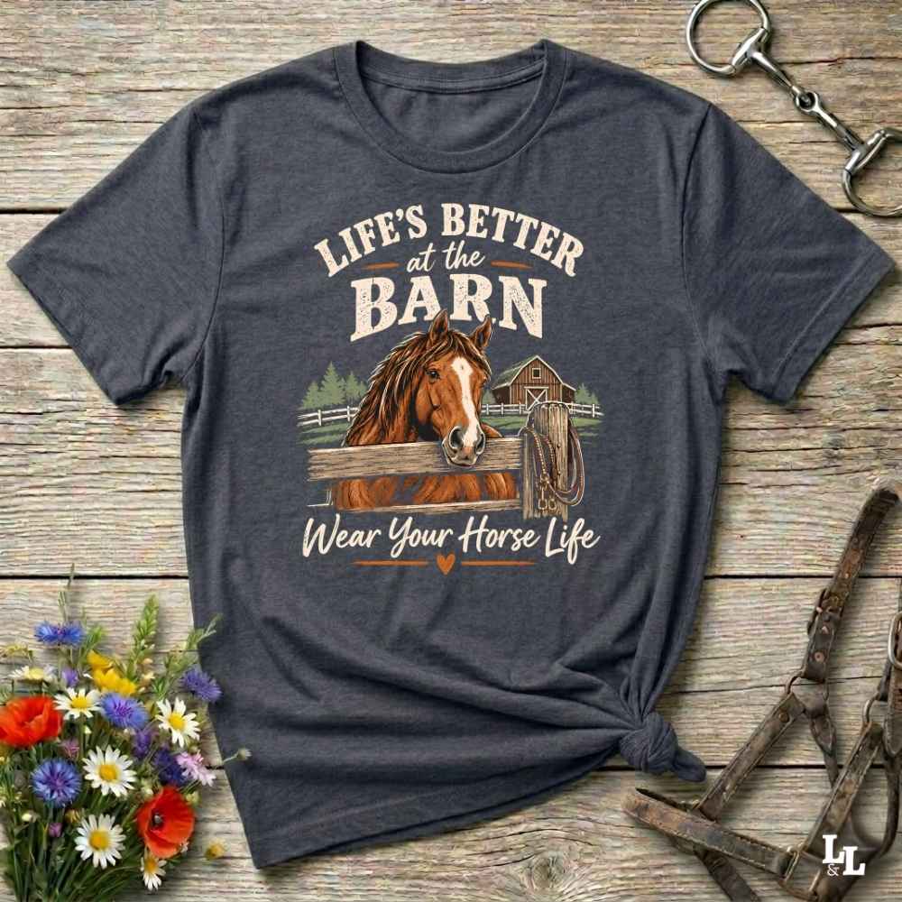 A dark heather gray graphic t-shirt featuring a brown horse and the text 'LIFE'S BETTER at the BARN Wear Your Horse Life' in a styled flat-lay on a rustic wooden background with flowers and horse tack.