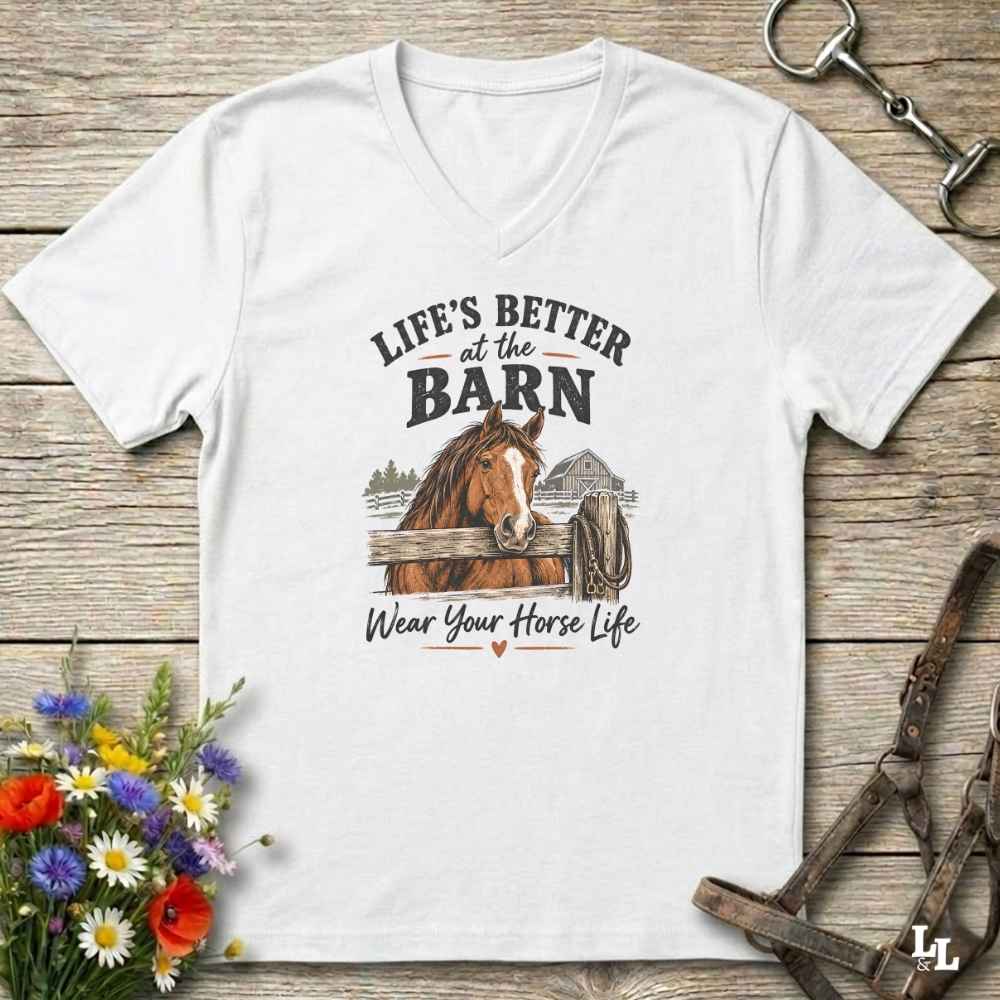 A white V-neck t-shirt with a graphic print of a brown horse and the text 'LIFE'S BETTER at the BARN Wear Your Horse Life', displayed on a rustic wooden background with equestrian accessories and flowers.