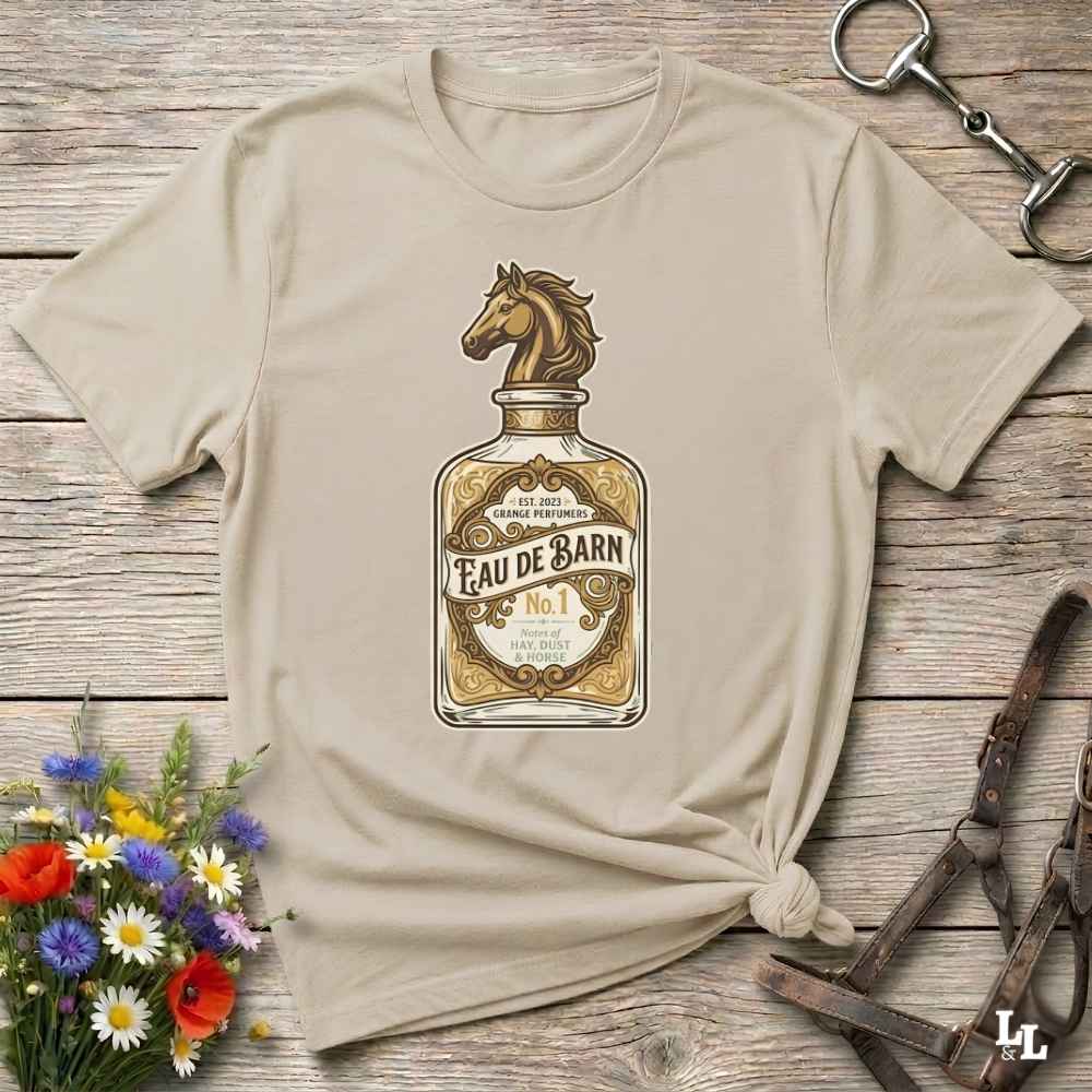 A beige short-sleeve t-shirt with a graphic of a horse-shaped bottle labeled 'Feau de Barn No. 1', styled flat on a rustic wooden background with equestrian accessories and a small flower arrangement.