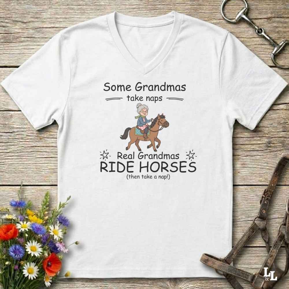 A white V-neck t-shirt with a graphic print of a grandma riding a horse and the text 'Some Grandmas take naps' and 'Real Grandmas RIDE HORSES (then take a nap!)'. The shirt is displayed flat on a rustic wooden background with equestrian-themed props like a leather bridle and flowers.