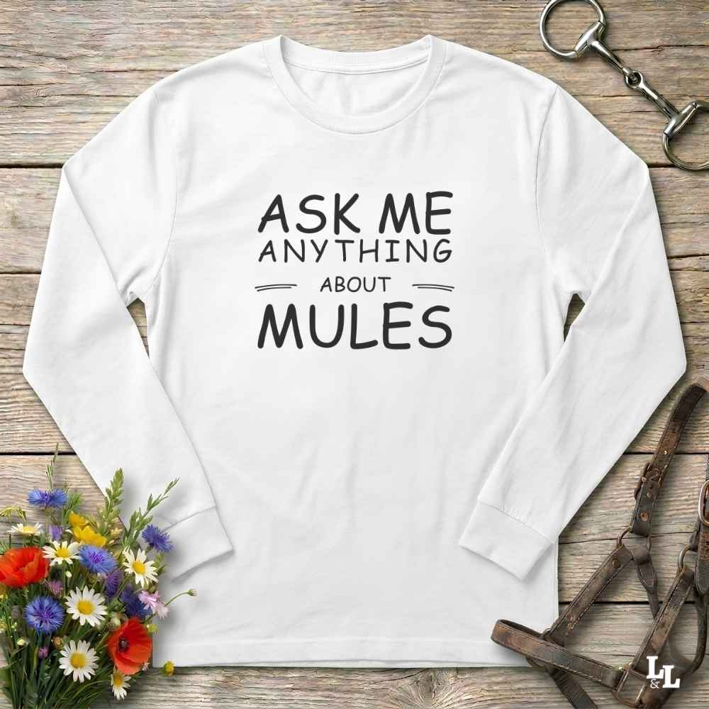 A white long-sleeve t-shirt with black text reading 'ASK ME ANYTHING ABOUT MULES' laid flat on a rustic wooden background with equestrian accessories and flowers.