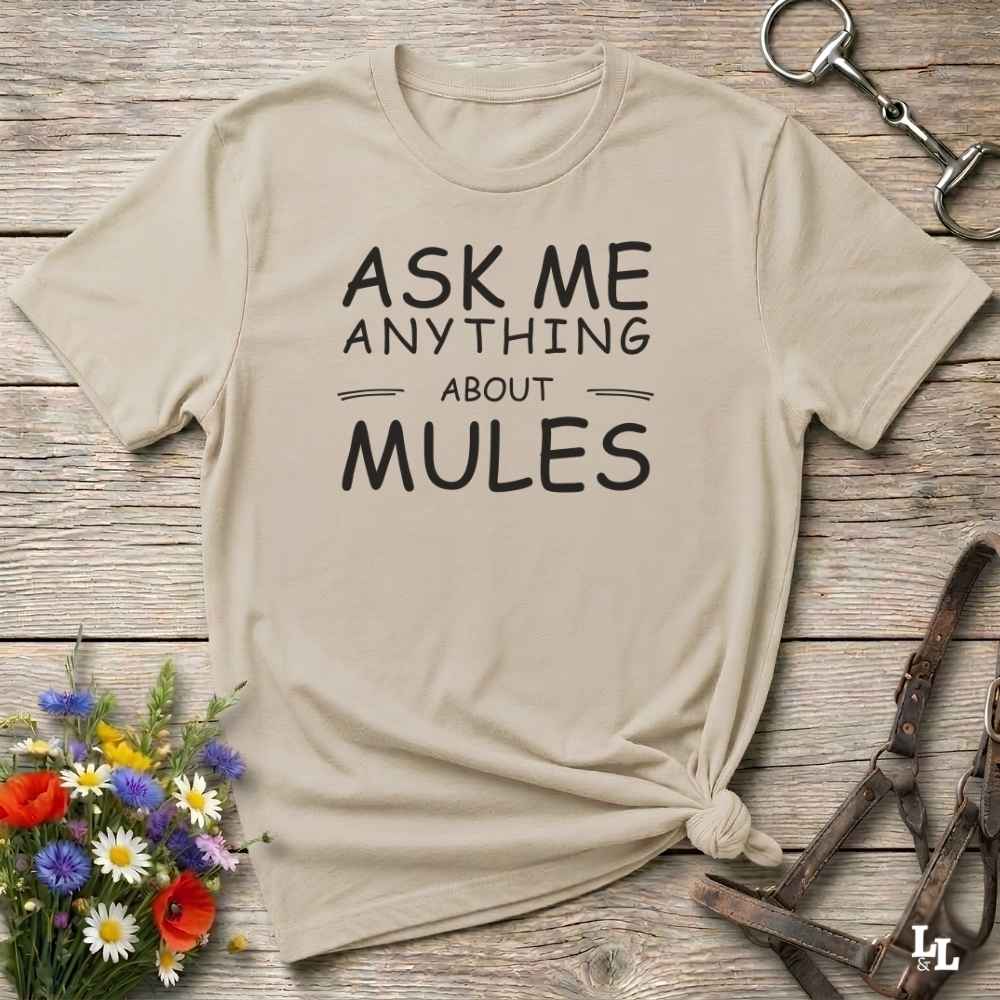 A beige graphic t-shirt with the text 'ASK ME ANYTHING ABOUT MULES' printed in black, styled in a flat-lay on a rustic wooden background with equestrian accessories and flowers.
