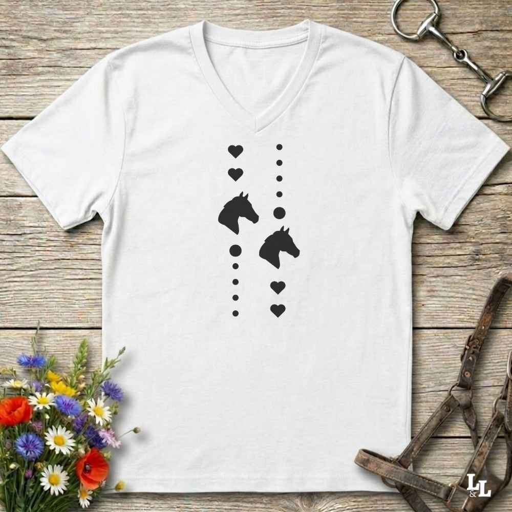 A white V-neck t-shirt with a black graphic print of horse heads and hearts, laid flat on a rustic wooden background with equestrian accessories and flowers.