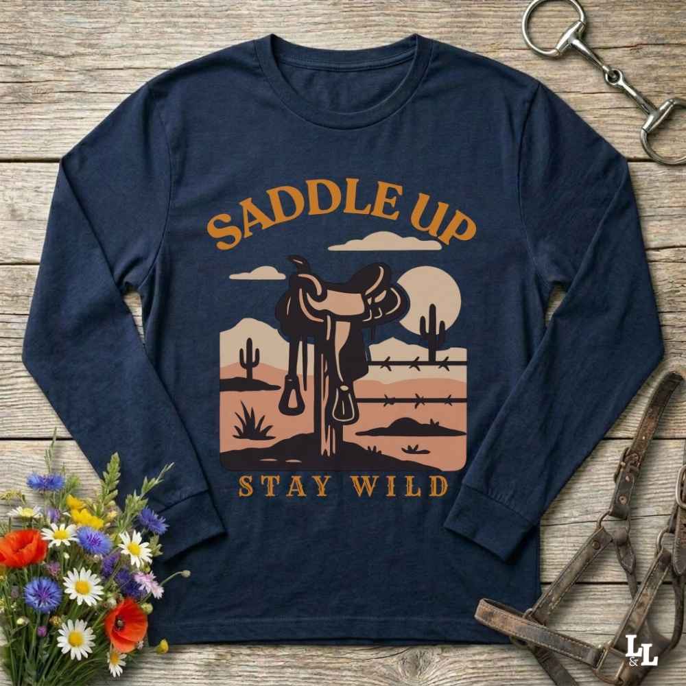 A navy blue long-sleeve t-shirt with a western-themed graphic print featuring a saddle, desert landscape, and the text 'SADDLE UP' and 'STAY WILD'. The shirt is displayed flat on a rustic wooden background with equestrian accessories and flowers.