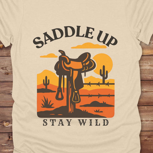 Saddle Up Stay Wild western t-shirt features a bold desert sunset graphic with a classic saddle and western landscape. Perfect for cowgirls, cowboys, and horse lovers, this rustic tee blends adventure with comfort and country charm. Whether you're heading to a rodeo, trail ride, or simply embracing your wild side, this shirt offers a soft fit and bold design. Designed for western wear fans, equestrians, and country souls, it's a must have for anyone who lives the saddle life.