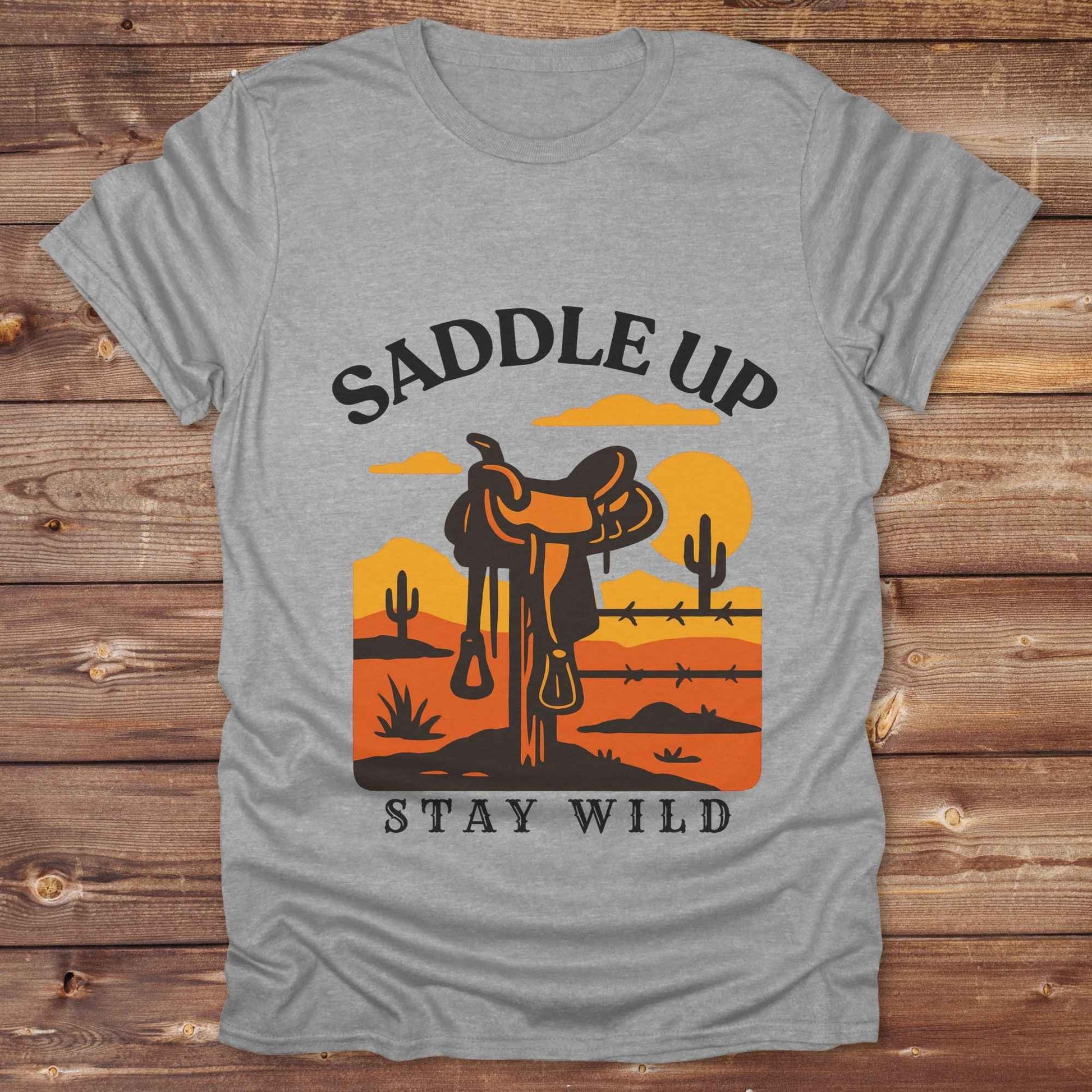 Saddle Up Stay Wild western t-shirt features a bold desert sunset graphic with a classic saddle and western landscape. Perfect for cowgirls, cowboys, and horse lovers, this rustic tee blends adventure with comfort and country charm. Whether you're heading to a rodeo, trail ride, or simply embracing your wild side, this shirt offers a soft fit and bold design. Designed for western wear fans, equestrians, and country souls, it's a must have for anyone who lives the saddle life.