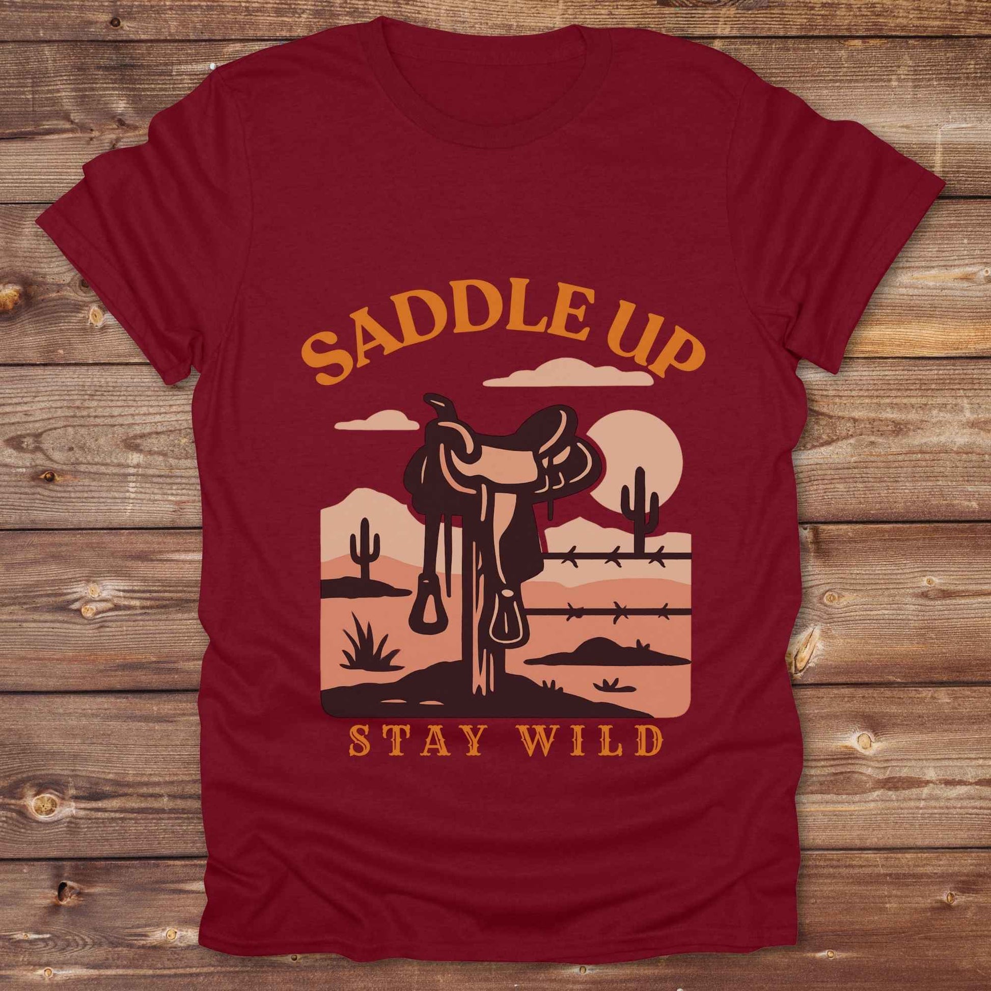 Saddle Up Stay Wild western t-shirt features a bold desert sunset graphic with a classic saddle and western landscape. Perfect for cowgirls, cowboys, and horse lovers, this rustic tee blends adventure with comfort and country charm. Whether you're heading to a rodeo, trail ride, or simply embracing your wild side, this shirt offers a soft fit and bold design. Designed for western wear fans, equestrians, and country souls, it's a must have for anyone who lives the saddle life.