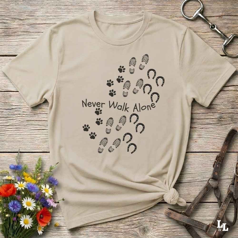 A beige short-sleeve t-shirt with a black graphic print of paw prints, hoof prints, and horseshoes around the text 'Never Walk Alone', styled flat on a rustic wooden background with flowers and horse tack props.