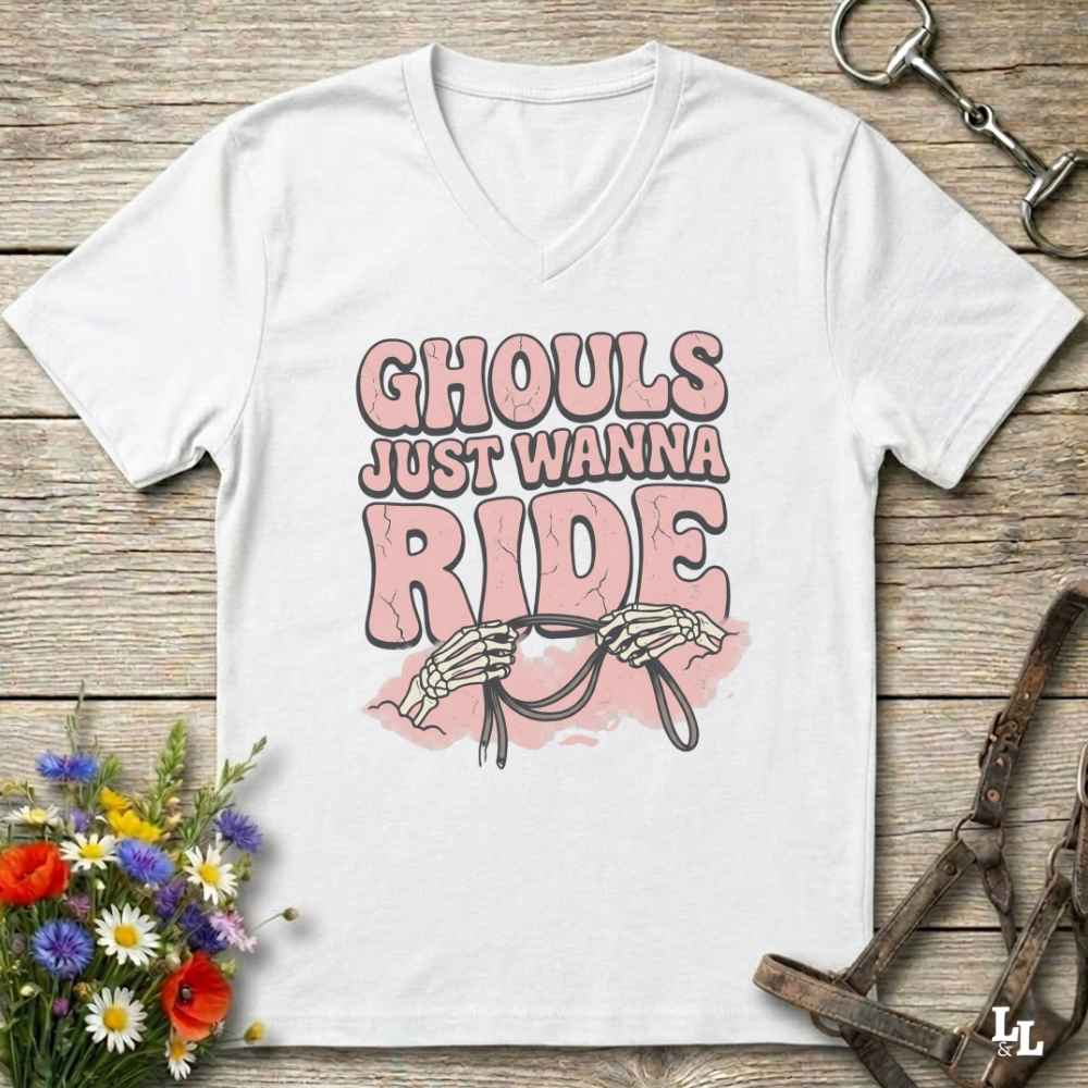A white V-neck t-shirt with a graphic print that reads 'GHOULS JUST WANNA RIDE' in pink and brown lettering, featuring skeleton hands holding reins. The shirt is displayed flat on a rustic wooden background with equestrian tack and flowers as props.