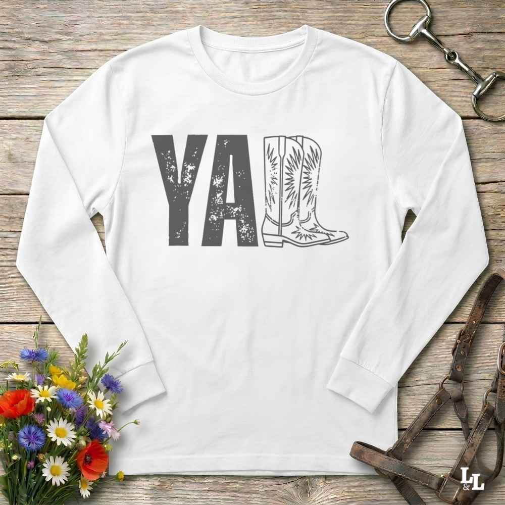 A white long-sleeve t-shirt with a graphic print reading 'Y'ALL' in distressed black letters, where the letters 'LL' are replaced by a western boot illustration. The shirt is laid flat on a rustic wooden surface with decorative props including a bouquet of flowers, a horse bit, and a leather bridle.