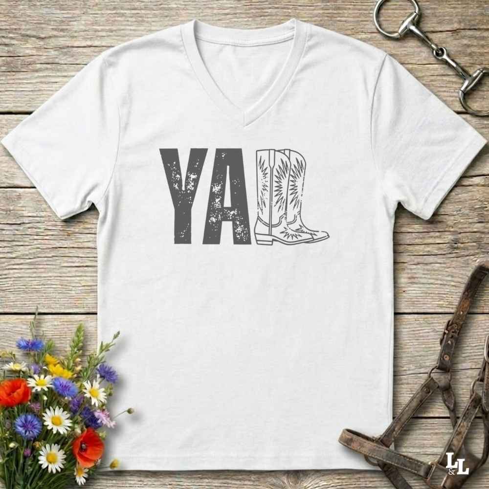 A white V-neck t-shirt with a western-themed graphic reading 'Y'ALL' in distressed black letters, where the letter 'L' is replaced by a cowboy boot illustration. The shirt is displayed flat on a rustic wooden background with equestrian tack and flowers.