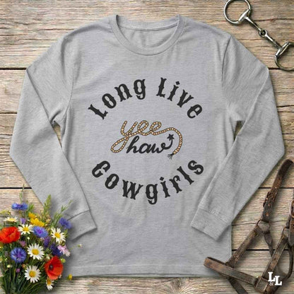 A light heather gray long-sleeve t-shirt with a graphic reading 'Long Live yee haw cowgirls' in black and glittery gold lettering, laid flat on a rustic wooden surface with decorative flowers and equestrian hardware around it.