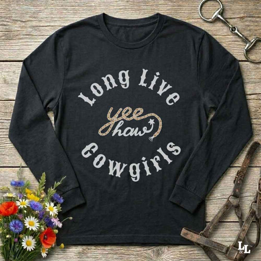 A black long-sleeve graphic t-shirt laid flat on a rustic wooden surface, surrounded by decorative props including a horse bit, a leather bridle, and a small bouquet of colorful wildflowers. The shirt features a graphic print that reads 'Long Live yeehaw Cowgirls' in white and glittery gold lettering.