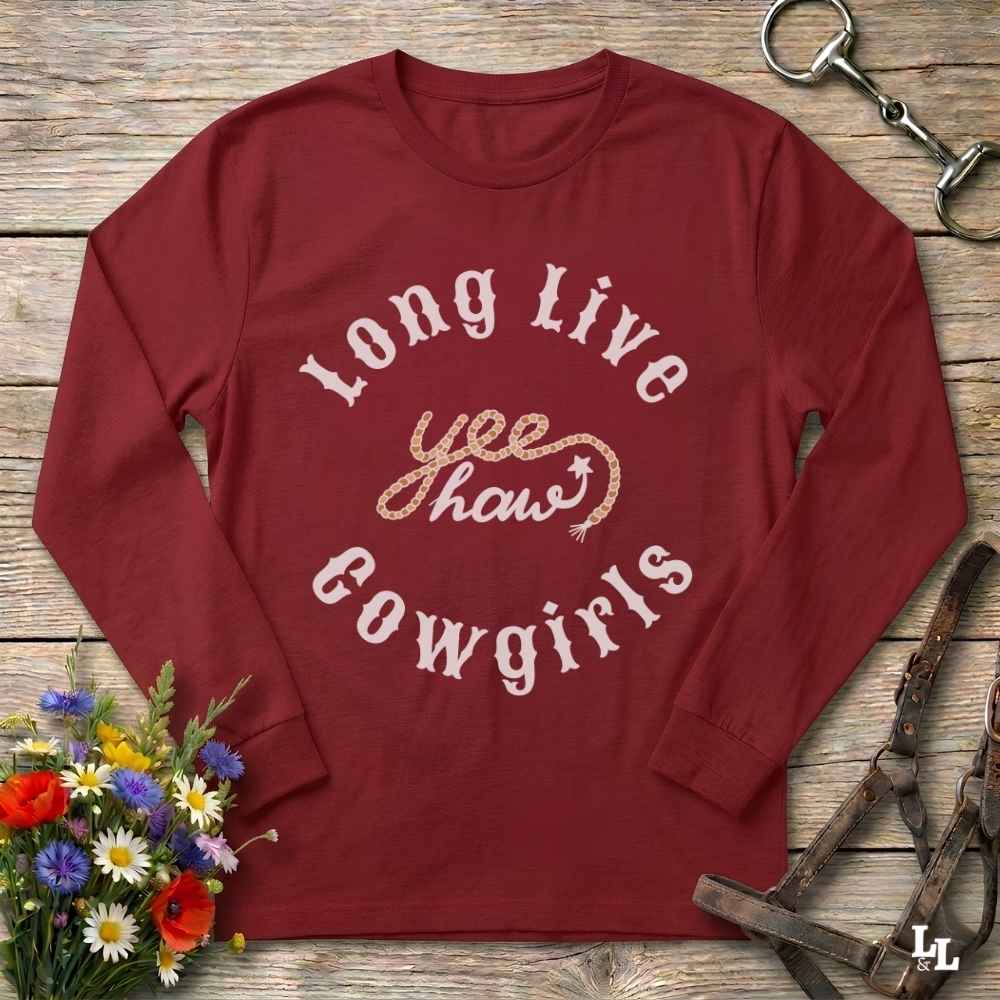 A long-sleeve crewneck t-shirt in maroon red, featuring a distressed white graphic that reads 'Long Live yeehaw Cowgirls' with a gold glittery 'yeehaw' in the center. The shirt is displayed flat on a rustic wooden background, styled with equestrian leather tack in the top right and a small bouquet of colorful wildflowers in the bottom left.
