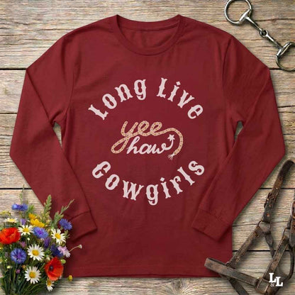 A long-sleeve crewneck t-shirt in maroon red, featuring a distressed white graphic that reads 'Long Live yeehaw Cowgirls' with a gold glittery 'yeehaw' in the center. The shirt is displayed flat on a rustic wooden background, styled with equestrian leather tack in the top right and a small bouquet of colorful wildflowers in the bottom left.