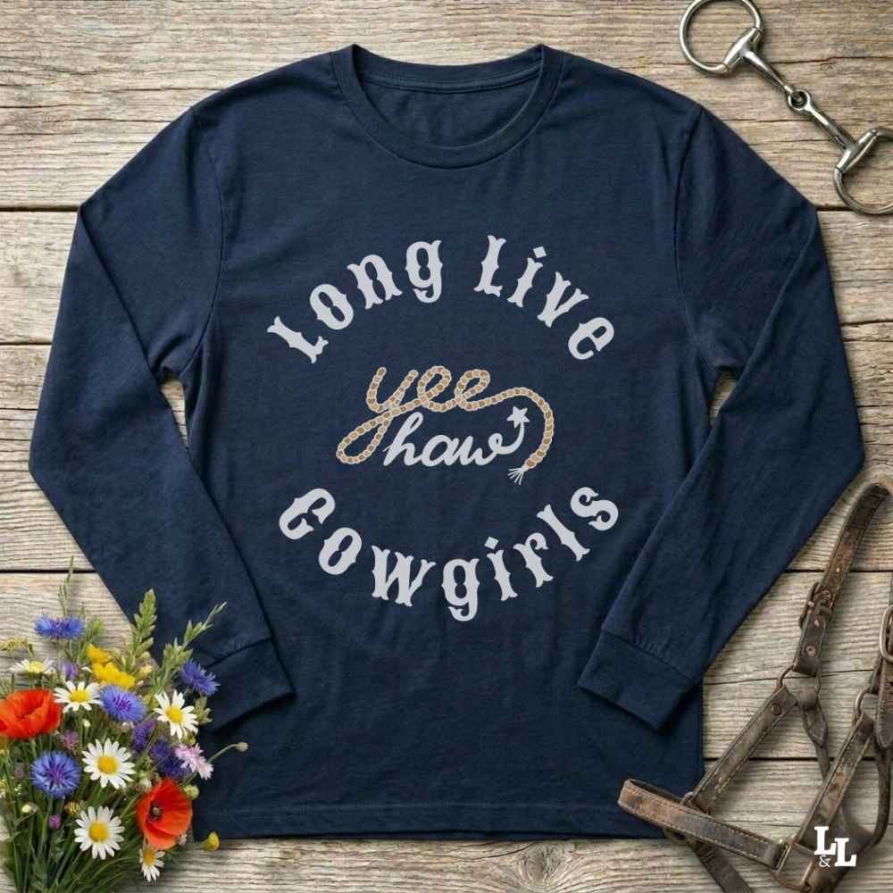 A navy blue long-sleeve graphic t-shirt laid flat on a rustic wooden surface, surrounded by cowboy-themed props including a halter, horse bit, and wildflowers. The shirt features a graphic print reading 'Long Live yeehaw Cowgirls' in white and glittery gold lettering.